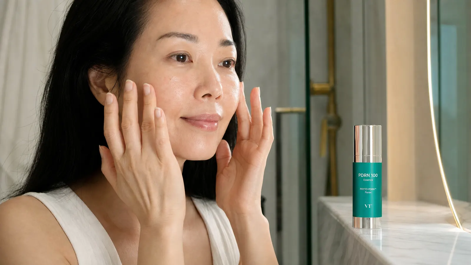 Hands pressing a few drops of clear PDRN essence into cheekbones, with a VT COSMETICS bottle visible in the frame