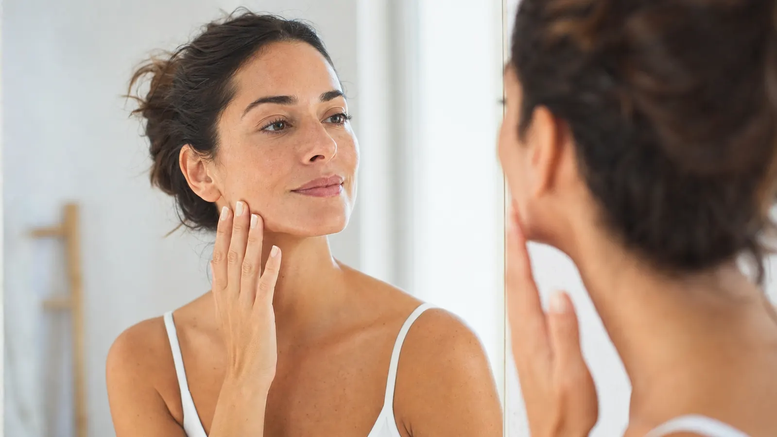 Woman with glowing skin examining her face in a well-lit bathroom mirror, looking calm and satisfied