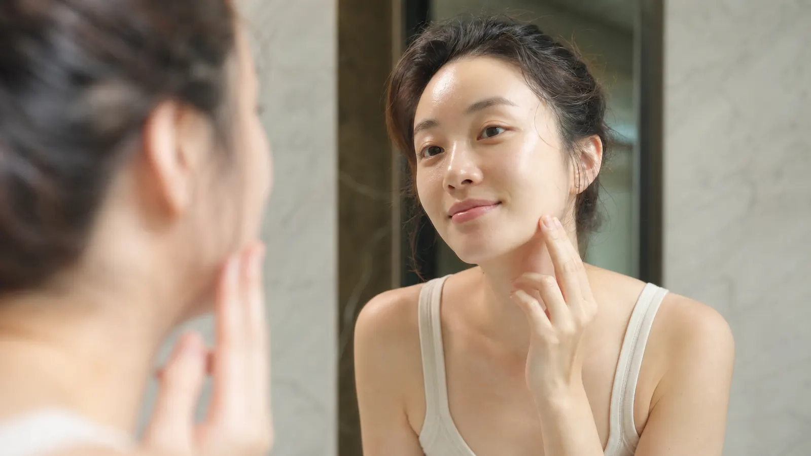 Woman with visibly smooth glowing skin looking into a mirror in a well-lit bathroom, suggesting the outcome of a consistent repair serum routine