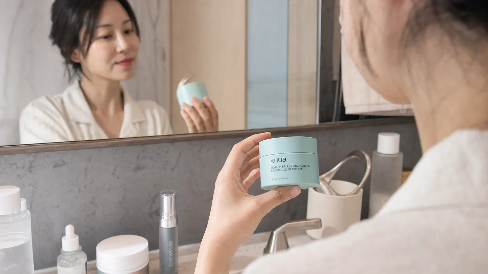 A hand pressing two fingers into the open lid of Anua PDRN Hyaluronic Acid 100 Moisturizing Cream, showing the white gel-cream texture